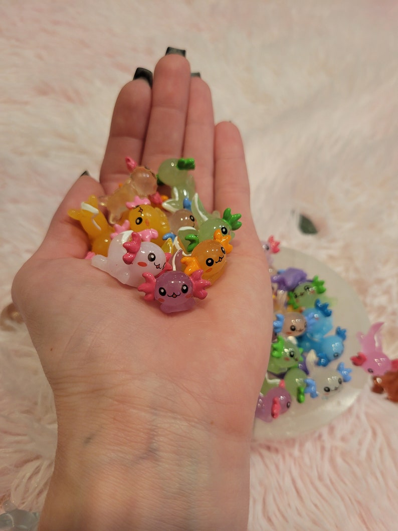 May include: A collection of colorful, small, axolotl figurines with smiling faces. The figurines are made of resin and have a glossy finish.