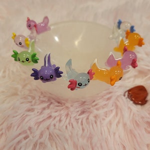May include: A collection of colorful axolotl figurines in a white bowl. The axolotls are made of glass and have a variety of colors, including purple, green, orange, and pink.