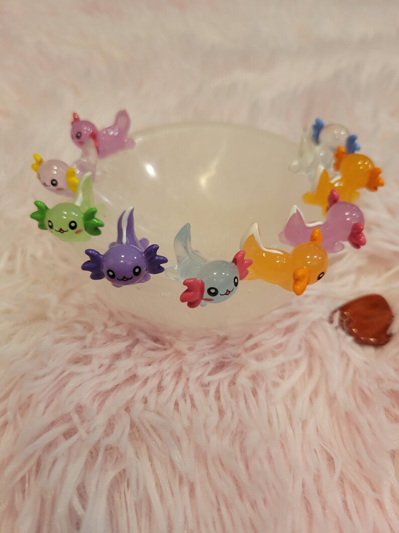 May include: A collection of colorful axolotl figurines in a white bowl. The axolotls are made of glass and have a variety of colors, including purple, green, orange, and pink.