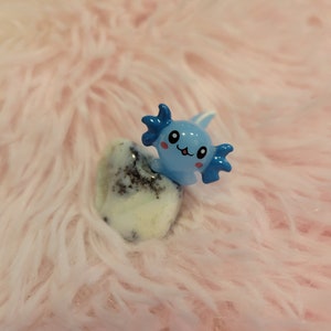 May include: A blue and white axolotl figurine with a smiling face, sitting on a white and black speckled stone. The figurine is on a pink fuzzy surface.