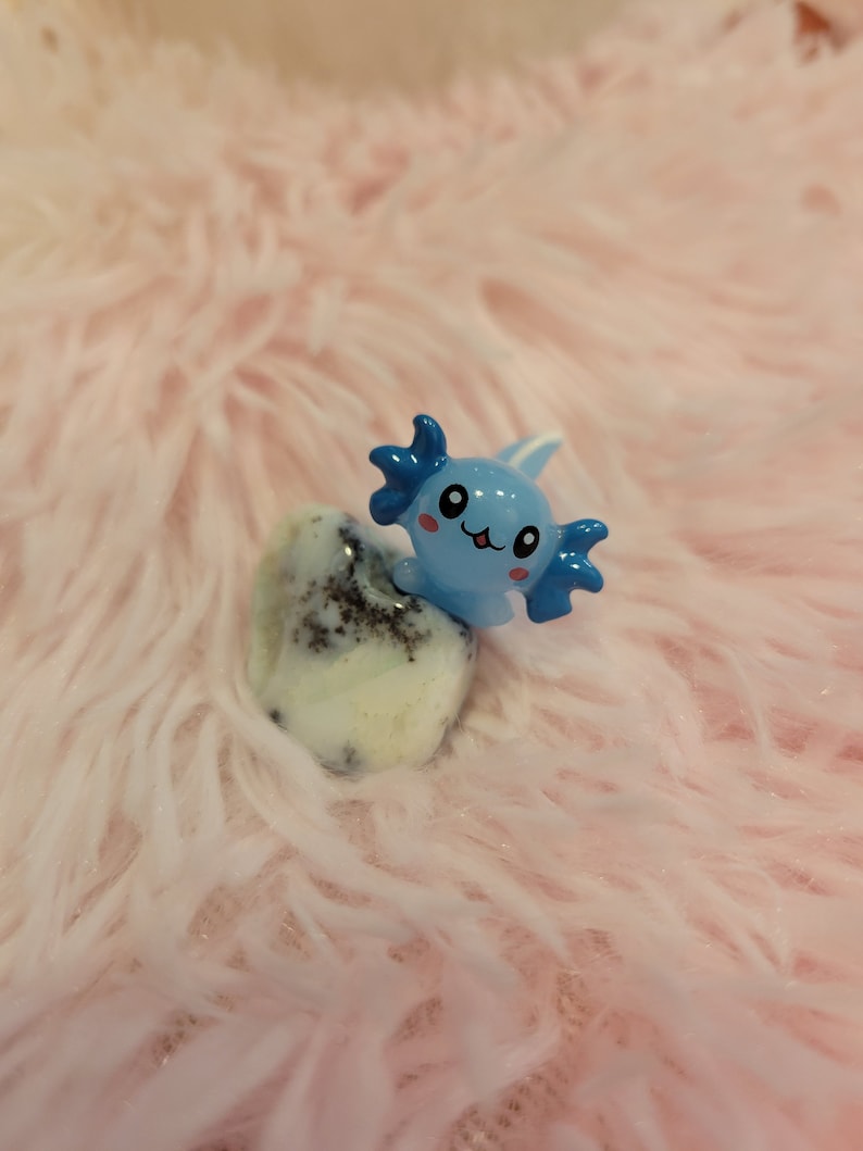 May include: A blue and white axolotl figurine with a smiling face, sitting on a white and black speckled stone. The figurine is on a pink fuzzy surface.