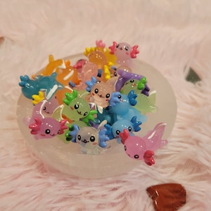 May include: A white bowl filled with colorful axolotl figurines. The axolotls are made of plastic and have different colors, including pink, blue, green, and yellow. The bowl is sitting on a pink fuzzy surface.