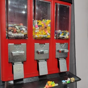 May include: A red gumball machine with three clear compartments filled with colorful items. The compartments hold polished stones, rubber ducks, and more stones. Each compartment has a "QUARTERS ONLY!" label. The machine has silver levers that say "THANK YOU".