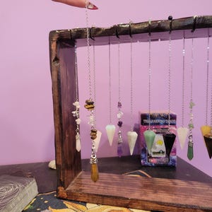 May include: A wooden display stand holds several crystal pendulums suspended by silver chains. The pendulums feature various gemstones and shapes, including teardrops and faceted points. A deck of tarot cards and a box of "The Learning Tarot" cards are also visible.