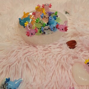 May include: A white bowl filled with colorful axolotl figurines. The axolotls are made of resin and have different expressions on their faces. The bowl is sitting on a pink fluffy surface.