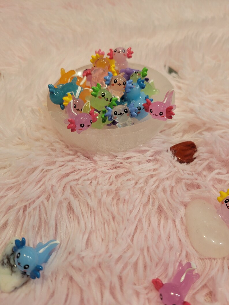 May include: A white bowl filled with colorful axolotl figurines. The axolotls are made of resin and have different expressions on their faces. The bowl is sitting on a pink fluffy surface.