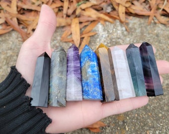 May include: A collection of eight crystal points in various colors, including black, grey, purple, blue, brown, white, green, and purple. The points are arranged in a row, held in a hand.