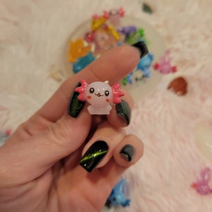 May include: A small pink and white axolotl figurine with black eyes and a happy face. The figurine is being held by a hand with black nail polish and green glitter.
