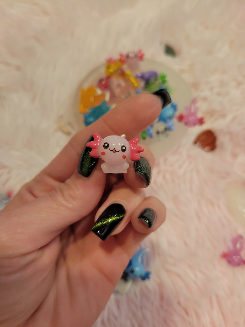 May include: A small pink and white axolotl figurine with black eyes and a happy face. The figurine is being held by a hand with black nail polish and green glitter.