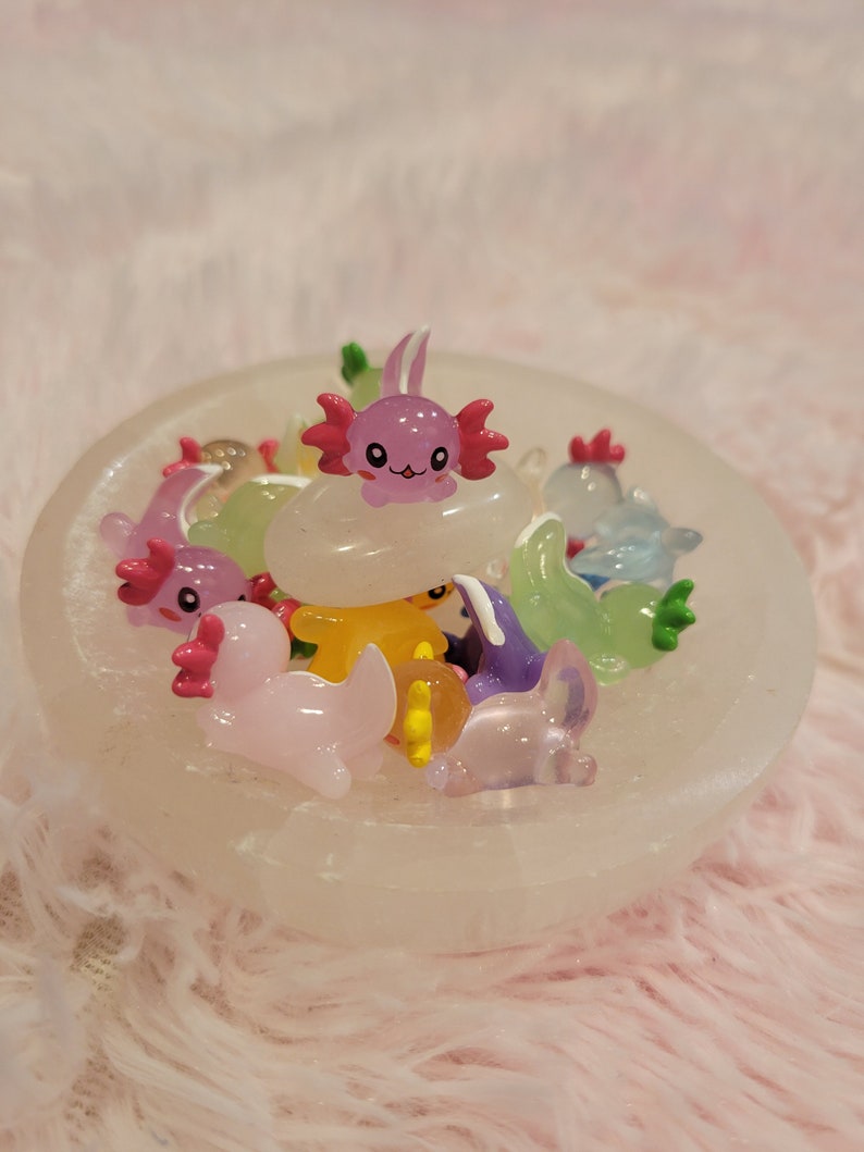 May include: A collection of colorful, small, plastic axolotl figurines in a white bowl. The axolotls are in various poses and have different colored eyes and fins.