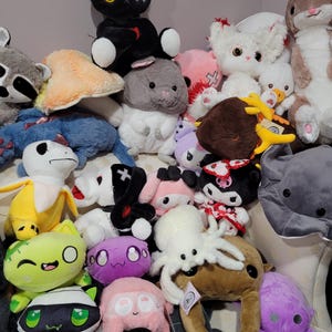 Pode incluir: Uma coleção de brinquedos de pelúcia em várias cores e designs, incluindo um gato preto, um coelho branco, um guaxinim marrom, um polvo rosa, um monstro roxo e um caranguejo marrom.