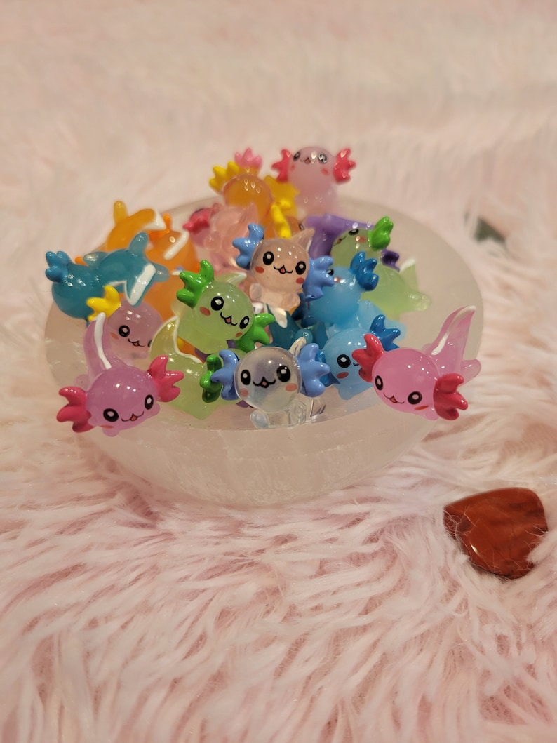 May include: A bowl filled with colorful, miniature axolotl figurines. The axolotls are made of a translucent plastic and have painted details, including eyes, gills, and smiles.