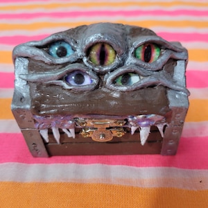 May include: A brown wooden chest with a silver trim and a collection of colorful eyes on top. The chest has a gold clasp and white teeth-like details along the bottom edge. The eyes are various colors, including blue, green, and red.