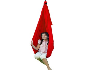 Pode incluir: Um balanço de tecido vermelho pendurado no teto com uma pessoa jovem sentada dentro. O balanço é feito de um tecido macio e elástico e é projetado para uso interno.