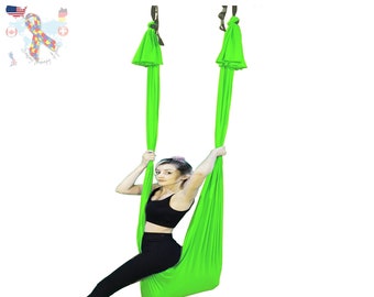Pode incluir: Um balanço de yoga verde pendurado no teto com uma pessoa sentada nele. O balanço é feito de tecido macio e é projetado para yoga aérea.