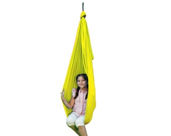 Pode incluir: Um balanço de tecido amarelo pendurado no teto com uma pessoa jovem sentada dentro. O balanço é feito de tecido macio e elástico e é projetado para uso interno ou externo.