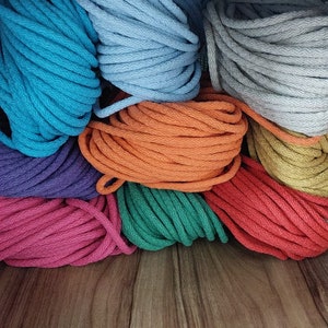 Pu&ograve; includere: Una collezione di corde di cotone colorate in varie tonalit&agrave; di blu, arancio, verde, rosso, viola e grigio. La corda &egrave; impilata ordinatamente su uno scaffale di legno.