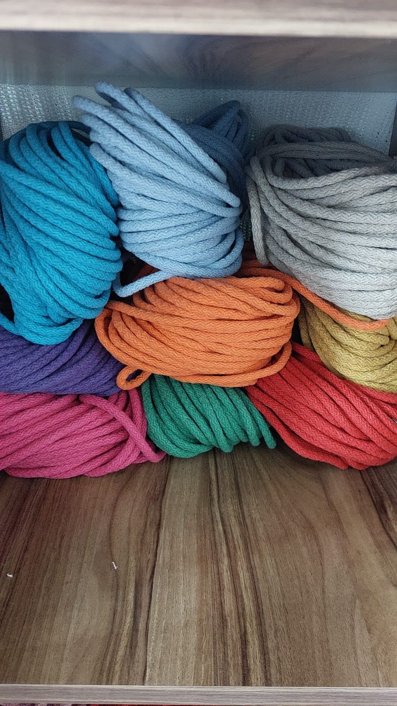 Pu&ograve; includere: Una collezione di corde di cotone colorate in varie tonalit&agrave; di blu, arancio, verde, rosso, viola e grigio. La corda &egrave; impilata ordinatamente su uno scaffale di legno.