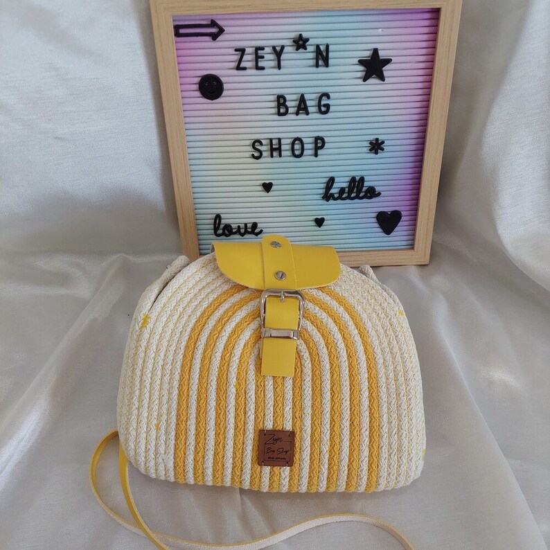 Pu&ograve; includere: Una borsa di paglia a righe gialle e bianche con una patta in pelle gialla e una fibbia. La borsa ha una tracolla lunga e un piccolo cartellino in pelle marrone con il testo "Zey'n Bag Shop" su di esso.