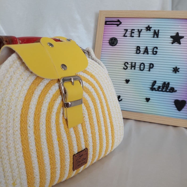Pu&ograve; includere: Borsa di paglia a righe gialle e bianche con una patta e una fibbia in pelle gialla. La borsa ha un'etichetta in pelle marrone con il testo "Zey'n Bag Shop" su di essa.