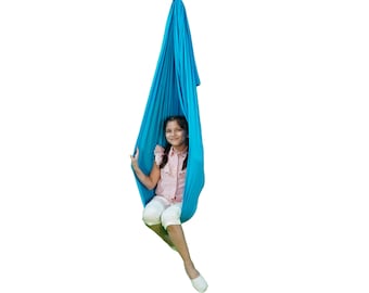 Pode incluir: Uma pessoa jovem está sentada em um balanço de tecido azul pendurado no teto. O balanço é feito de tecido macio e elástico e é projetado para uso interno.