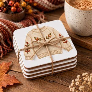 Könnte beinhalten: Ein Stapel aus vier quadratischen, weißen Untersetzern, die mit braunem Bindfaden zusammengebunden sind. Jeder Untersetzer zeigt eine Illustration eines beigen Strickpullovers mit gestickten Herbstblättern und Beeren in Orange, Rot und Grün.