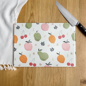 May include: Rectangular glass cutting board with a white background and a colorful fruit pattern. The pattern includes pink apples, green pears, orange citrus fruits, and red cherries. A stainless steel knife with a black handle rests on the wooden surface.