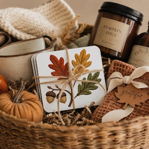 Puede incluir: Una cesta de regalo con un conjunto de posavasos cuadrados atados con cuerda, con una ilustración de hojas de roble rojas, amarillas y verdes con bellotas. Incluye una vela Pumpkin Spice, una pequeña calabaza de tela naranja y un paño de tejido gofrado.