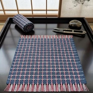 May include: A rectangular woven textile mat featuring a blue base with a grid pattern of thin white and pink lines. The mat has a fringed edge with alternating pink and white threads. A matching rolled fabric piece and metal scissors are nearby.