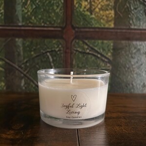 May include: A clear glass candle holder with a white soy candle. The candle has a white wick and a label that reads "Joyful Light Living Soy Candles" with a heart symbol. The candle sits on a dark wooden surface.