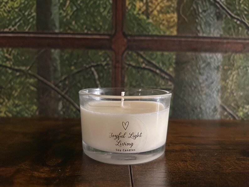 May include: A clear glass candle holder with a white soy candle. The candle has a white wick and a label that reads "Joyful Light Living Soy Candles" with a heart symbol. The candle sits on a dark wooden surface.