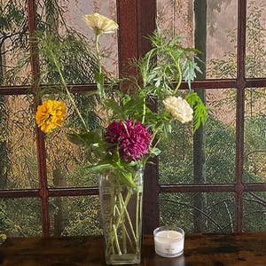May include: A vase of flowers with yellow, white, and magenta blooms. A small candle in a glass container is on a dark wooden surface. The background is a window with a forest scene. The vase is clear glass.
