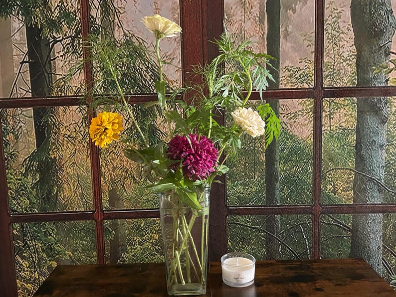 May include: A vase of flowers with yellow, white, and magenta blooms. A small candle in a glass container is on a dark wooden surface. The background is a window with a forest scene. The vase is clear glass.