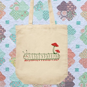 Puede incluir: Bolso tote de lona beige con una ilustraci&oacute;n de una oruga sonriente con pies rojos y setas rojas. La oruga tiene hierba verde en la espalda. El bolso tiene dos asas beige.