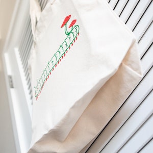 Puede incluir: Bolso tote de lona color crema con un dise&ntilde;o ilustrado en verde y rojo de una oruga y setas. El bolso cuelga sobre un fondo blanco con lamas. El bolso tote tiene un dise&ntilde;o sencillo y minimalista.