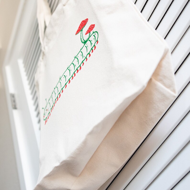 Puede incluir: Bolso tote de lona color crema con un dise&ntilde;o ilustrado en verde y rojo de una oruga y setas. El bolso cuelga sobre un fondo blanco con lamas. El bolso tote tiene un dise&ntilde;o sencillo y minimalista.