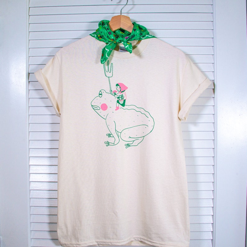 May include: Cream-coloured t-shirt featuring a green illustration of a person riding a frog, holding a fork. The frog has pink cheeks. A green bandana is draped over the hanger. The shirt has rolled-up sleeves.
