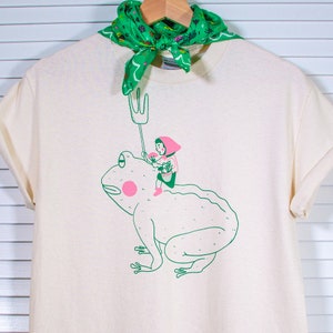 May include: A cream-coloured t-shirt with a quirky illustration of a person riding a frog, holding a pitchfork. The frog is green with a pink cheek. A green bandana with a floral pattern is draped over the shirt's neckline.