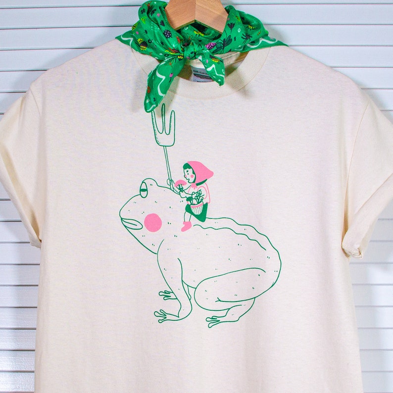 May include: A cream-coloured t-shirt with a quirky illustration of a person riding a frog, holding a pitchfork. The frog is green with a pink cheek. A green bandana with a floral pattern is draped over the shirt's neckline.