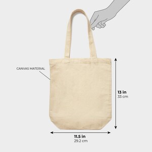 Puede incluir: Un bolso tote de lona beige, de 33 cm de alto y 29,2 cm de ancho, con dos asas a juego. La bolsa est&aacute; hecha de material de lona y la sostiene una mano. Las dimensiones tambi&eacute;n se muestran en pulgadas.