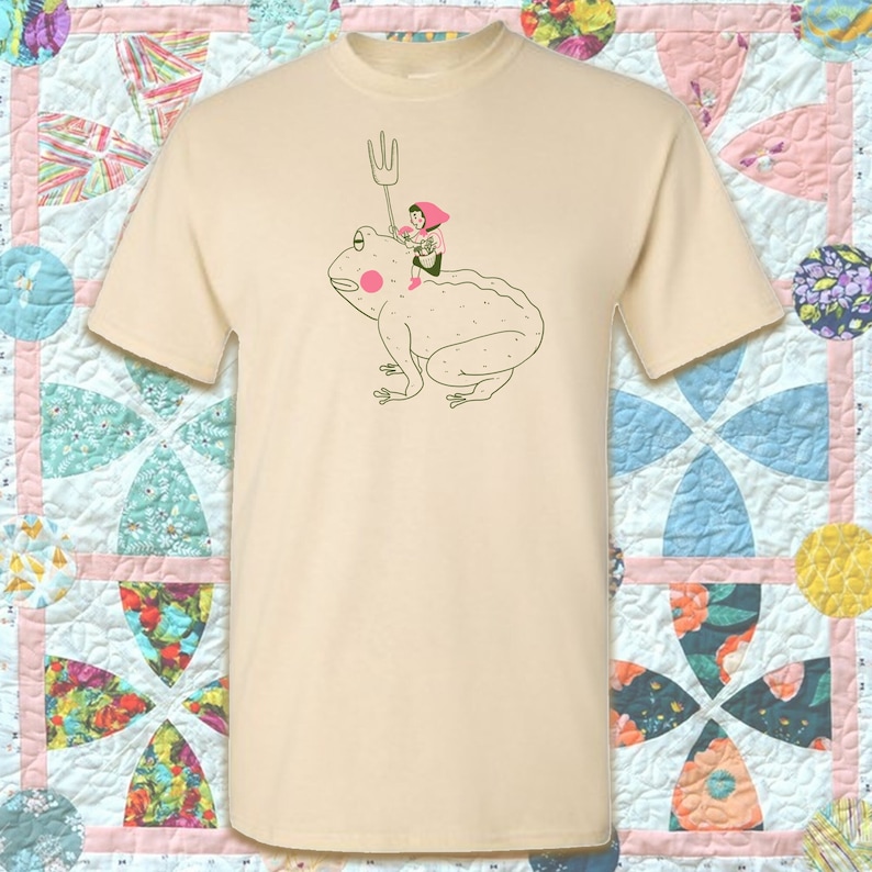 May include: A cream-coloured t-shirt with a quirky illustration. A person wearing a pink hat and holding a pitchfork is riding a large frog. The frog has a pink cheek and is outlined in green. The t-shirt is a casual, unisex style.