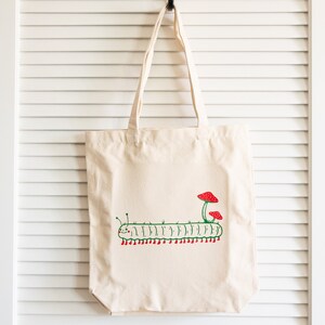 Puede incluir: Un bolso tote de color crema con un dise&ntilde;o caprichoso de oruga. La oruga verde tiene pies rojos y est&aacute; rematada con dos setas rojas. El bolso tiene asas largas para llevar al hombro.