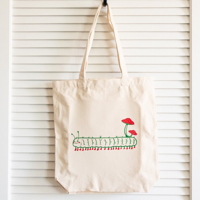 Puede incluir: Un bolso tote de color crema con un dise&ntilde;o caprichoso de oruga. La oruga verde tiene pies rojos y est&aacute; rematada con dos setas rojas. El bolso tiene asas largas para llevar al hombro.