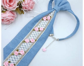 Könnte beinhalten: Eine hellblaue Jeans-Krawatte mit einem zentralen vertikalen Einsatz aus weißer Spitzenborte. Die Spitze ist von einer dünnen silbernen Metallpaspel umrandet und mit kleinen rosa Stoffrosen verziert. Die Krawatte hat eine verstellbare silberne Kette.