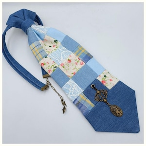 Può includere: Una cravatta patchwork con tessuti in denim e floreali nei toni del blu, crema e giallo. La cravatta è adornata da un ciondolo e una catena decorativi color bronzo. Un accessorio unico.