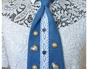 Könnte beinhalten: Eine blaue Jeans-Krawatte mit weißer Spitzenborte und dekorativen Herz- und Perlenakzenten. Die Krawatte ist mit goldfarbenen Verzierungen und kleinen blauen Herzdetails versehen. Die Krawatte wird über einem weißen Hemd mit Kragen und einem weißen Spitzentop getragen.