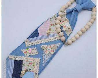 May include: A handcrafted denim tie with patchwork details, adorned with lace, floral fabric, and wooden beads. The tie features a light blue denim base with various fabric patches and decorative elements.