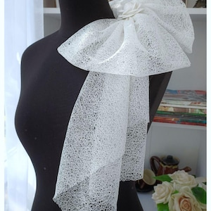 Op de afbeelding: Een witte, kanten sjaal of halsstuk, gedrapeerd over een zwarte mannequin. De sjaal heeft een grote strik aan de bovenkant en lange, vloeiende panelen. De delicate, open weefsel van de stof creëert een lichte en luchtige uitstraling.