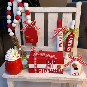 May include: A white wooden bench with red and white gingham ribbon, a red and white beaded garland, a red and white mug with whipped cream, a red and white box with the text "Fresh Sweet Strawberries", two red and white rolling pins with the text "Strawberry" and "Berry Sweet", and a white birdhouse with the text "Sweet".