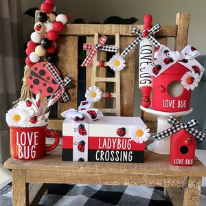 May include: A collection of ladybug-themed decorations, including a red mug with "LOVE BUG" text, a birdhouse, and a stack of books with "LADYBUG CROSSING" text. The decorations feature red, black, and white colors, with daisy accents and checkered ribbons.